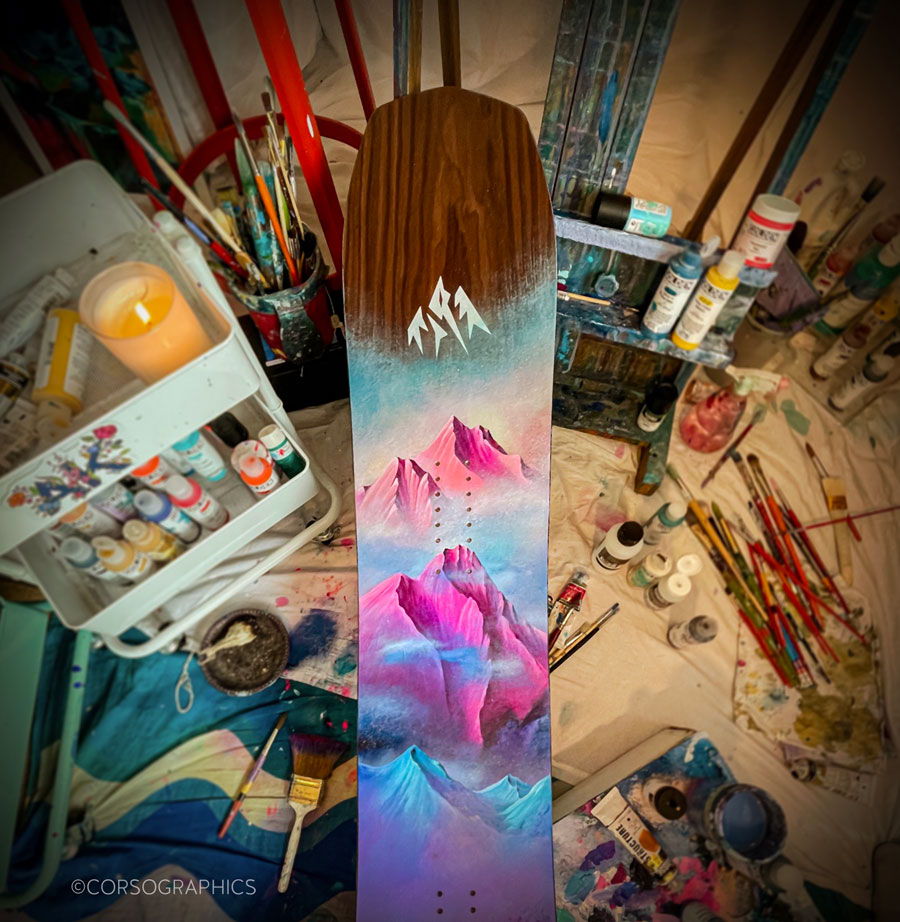 Colorful painted snowboard surrounded by art supplies and brushes.