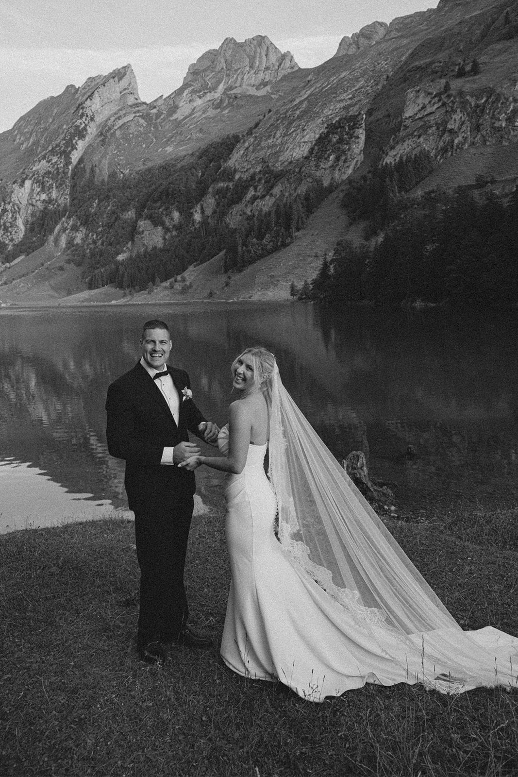 Bride and groom smiling by a lake with mountains in the background.