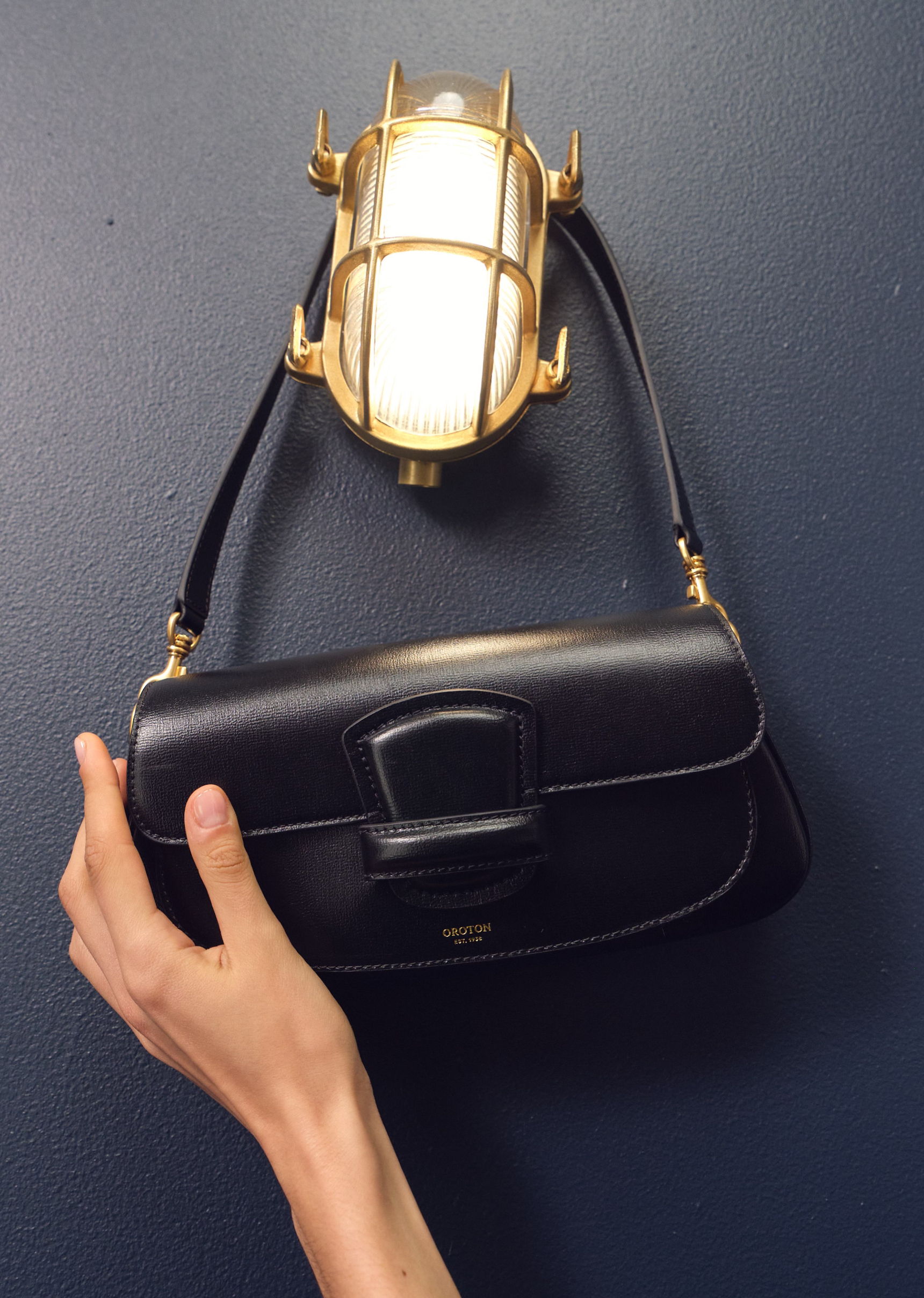 A hand holding a black handbag against a blue wall.
