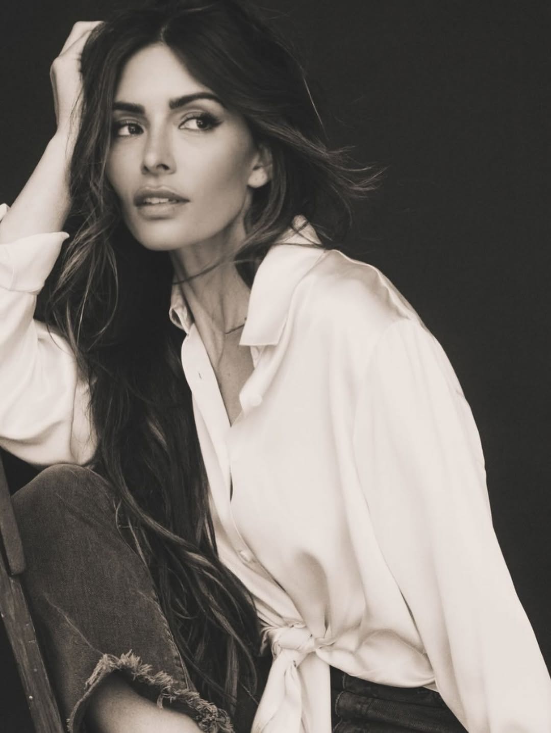 A woman with long hair wearing a white blouse, sitting in a relaxed pose.