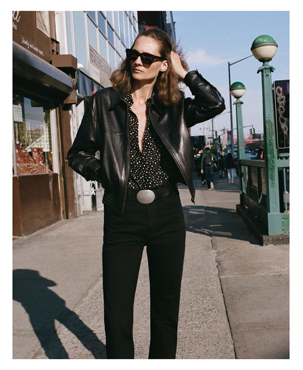 Woman in a black leather jacket and sunglasses, standing on a city street.