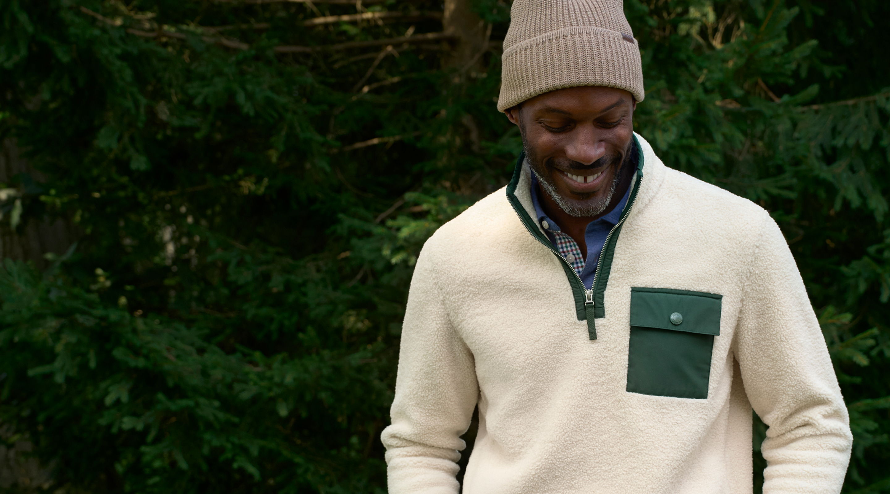 Smiling person wearing a sweater and a beanie in a forest setting.