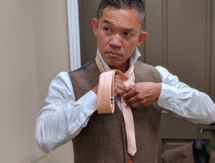 Man wearing a tan vest and tying a light pink necktie 