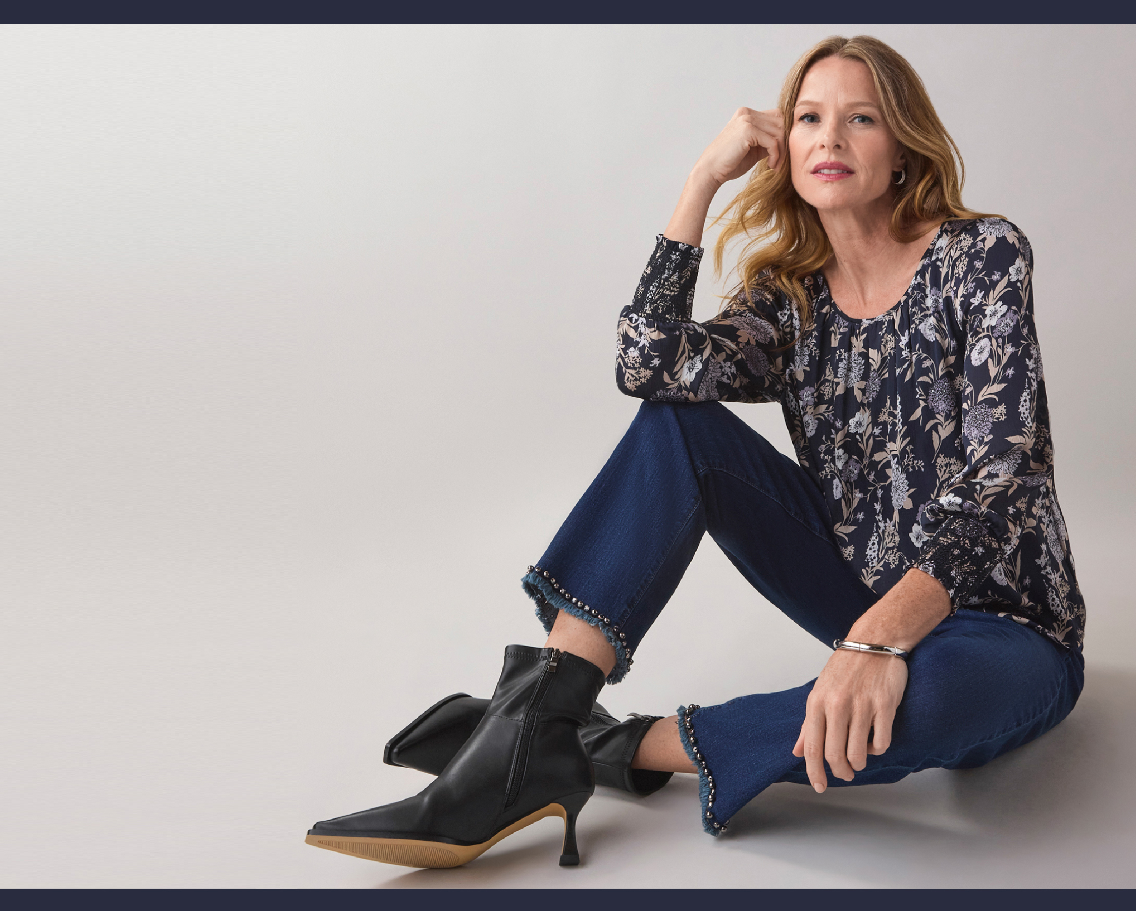 Woman sitting stylishly, wearing a patterned top and high-heeled boots.