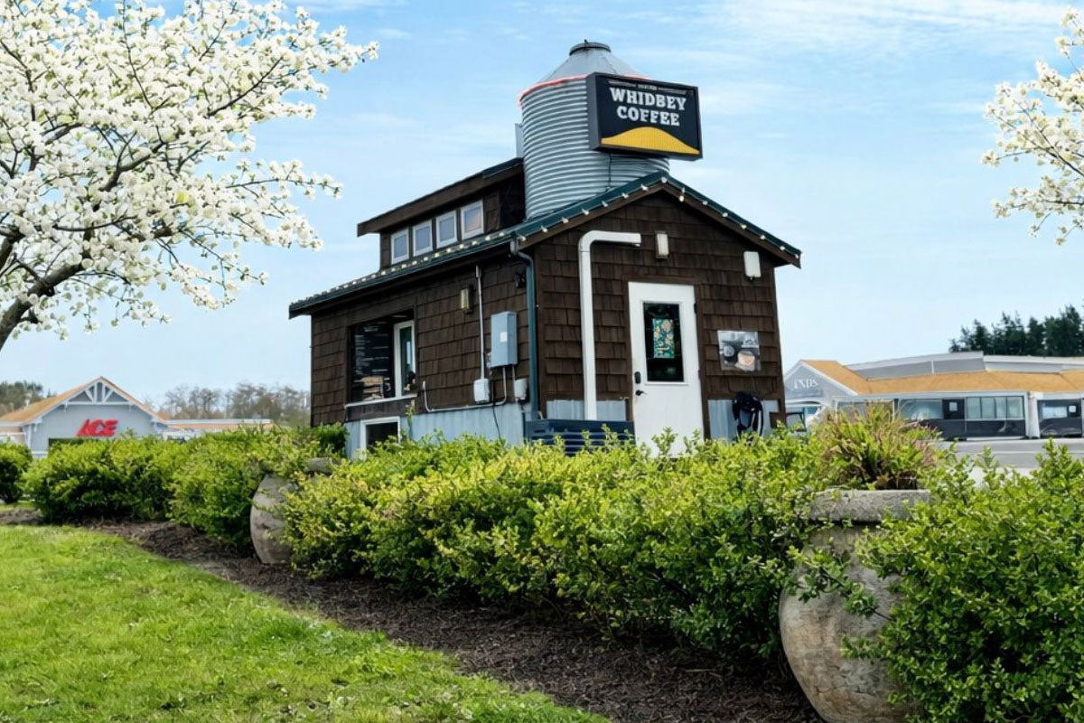 Whidbey Coffee building with a silo-like structure, surrounded by greenery and trees with white blossoms in Freeland, Washington.