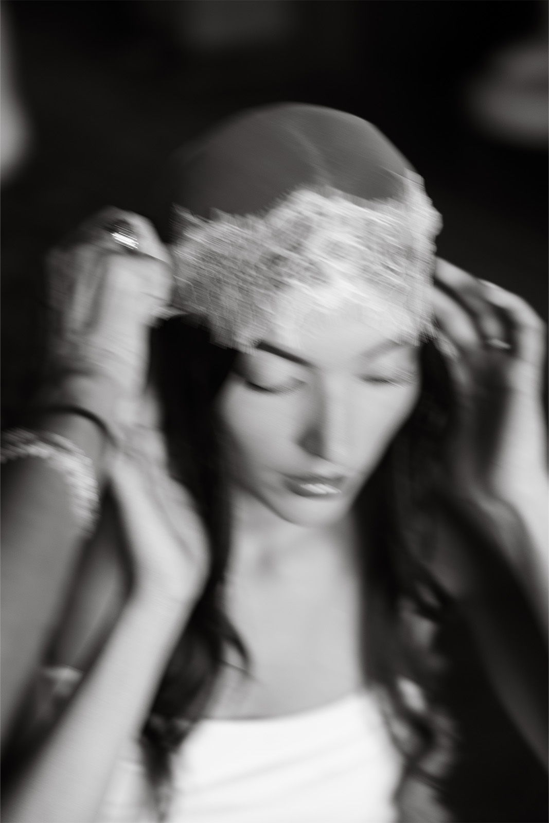 Woman in a white dress adjusting a lace headpiece, captured in black and white.