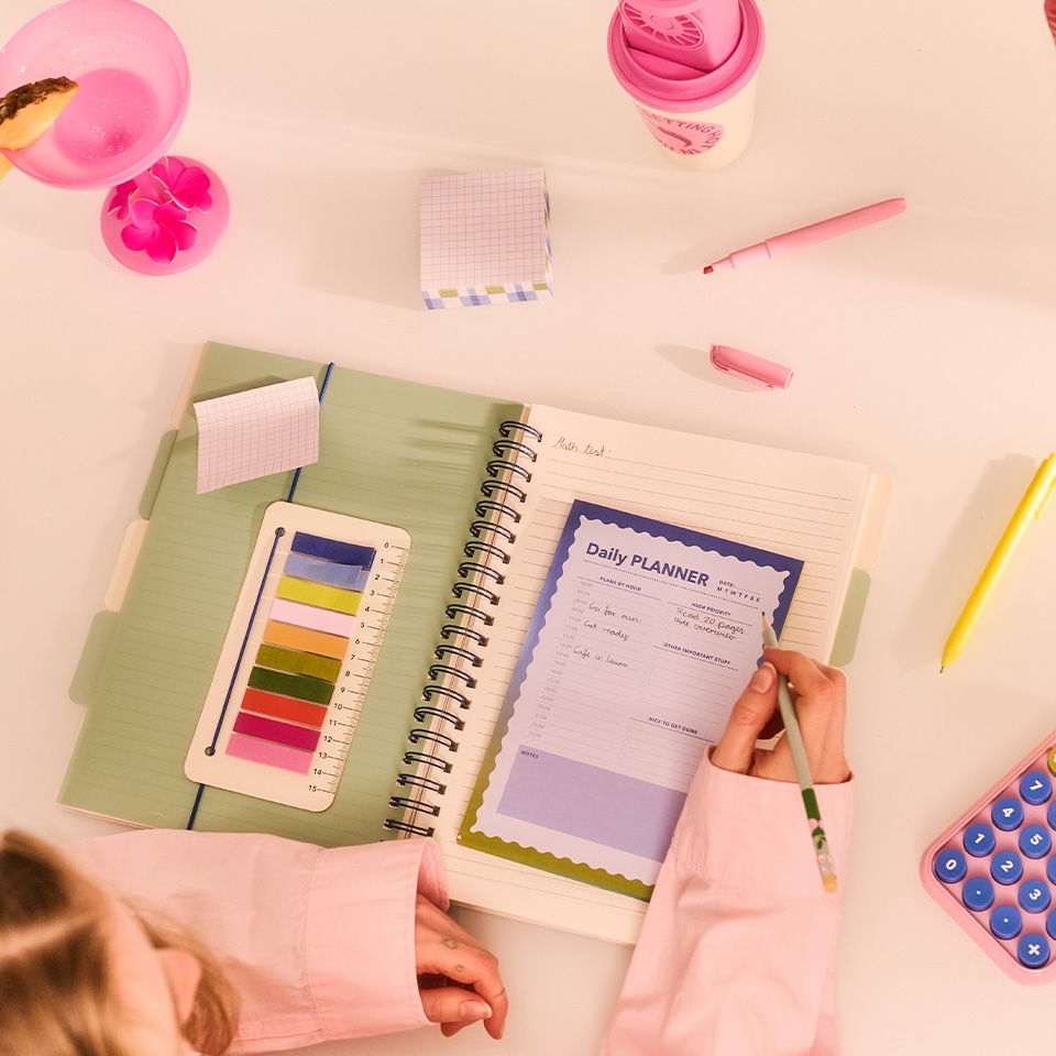 Person writing in a notebook with colorful stationery on a white desk.