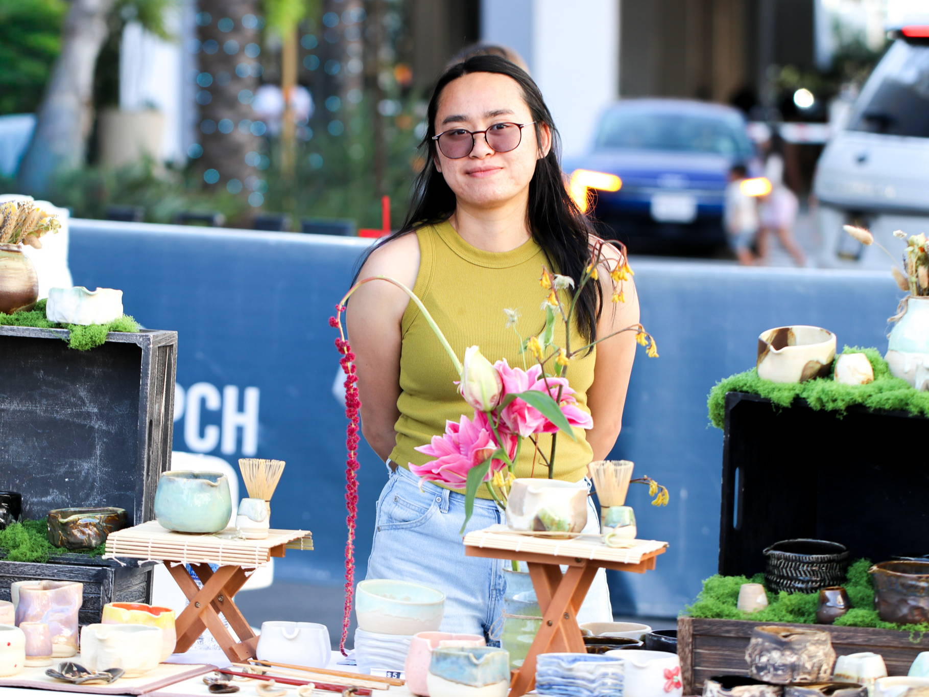 Sophia, owner of Sojha Studio, with flowers and ceramics at a market stall, wearing sunglasses and a yellow top.
