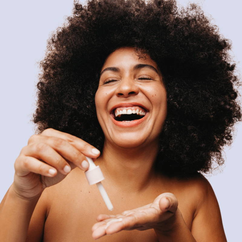 A smiling woman dropping a clear serum into her palm from an eye dropper