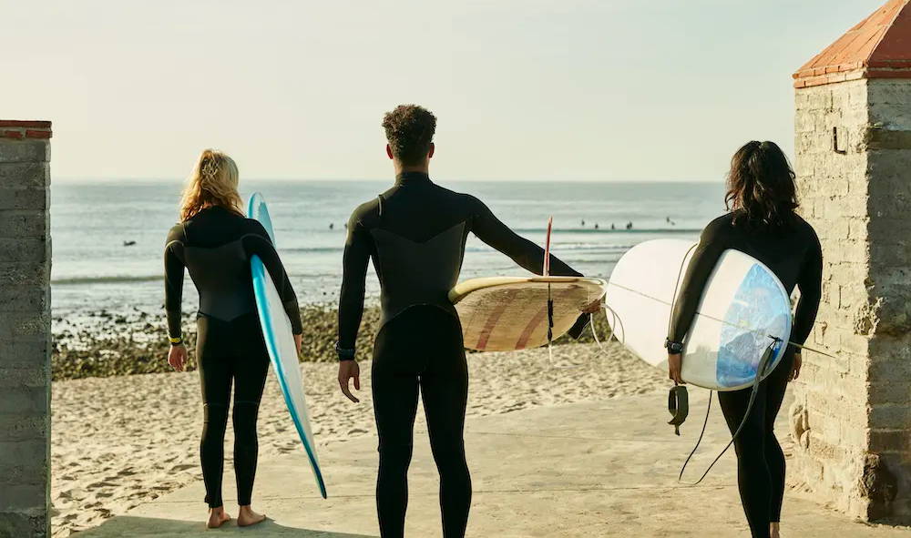 Three surfers with boards standing on the beach wearing Garmin Instinct 2 surf watches