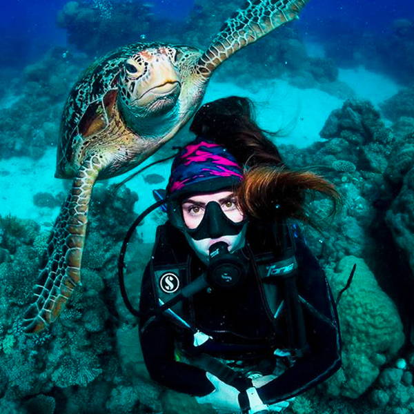 Sally Higgs in scuba gear, wearing a face shield as a headband, posing with a sea turtle.