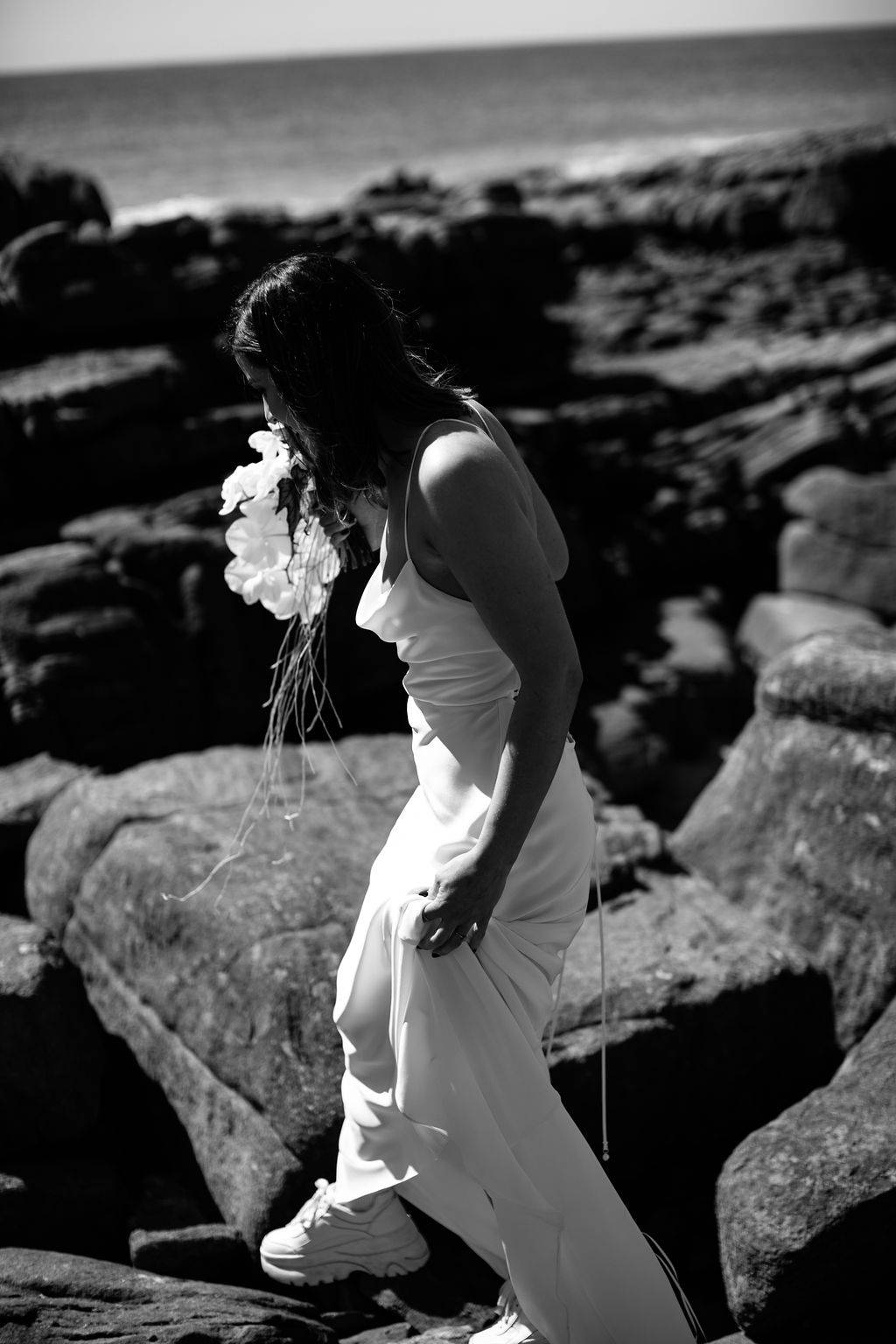 Bride wearing a silk white dress and white sneakers walking on the rocks