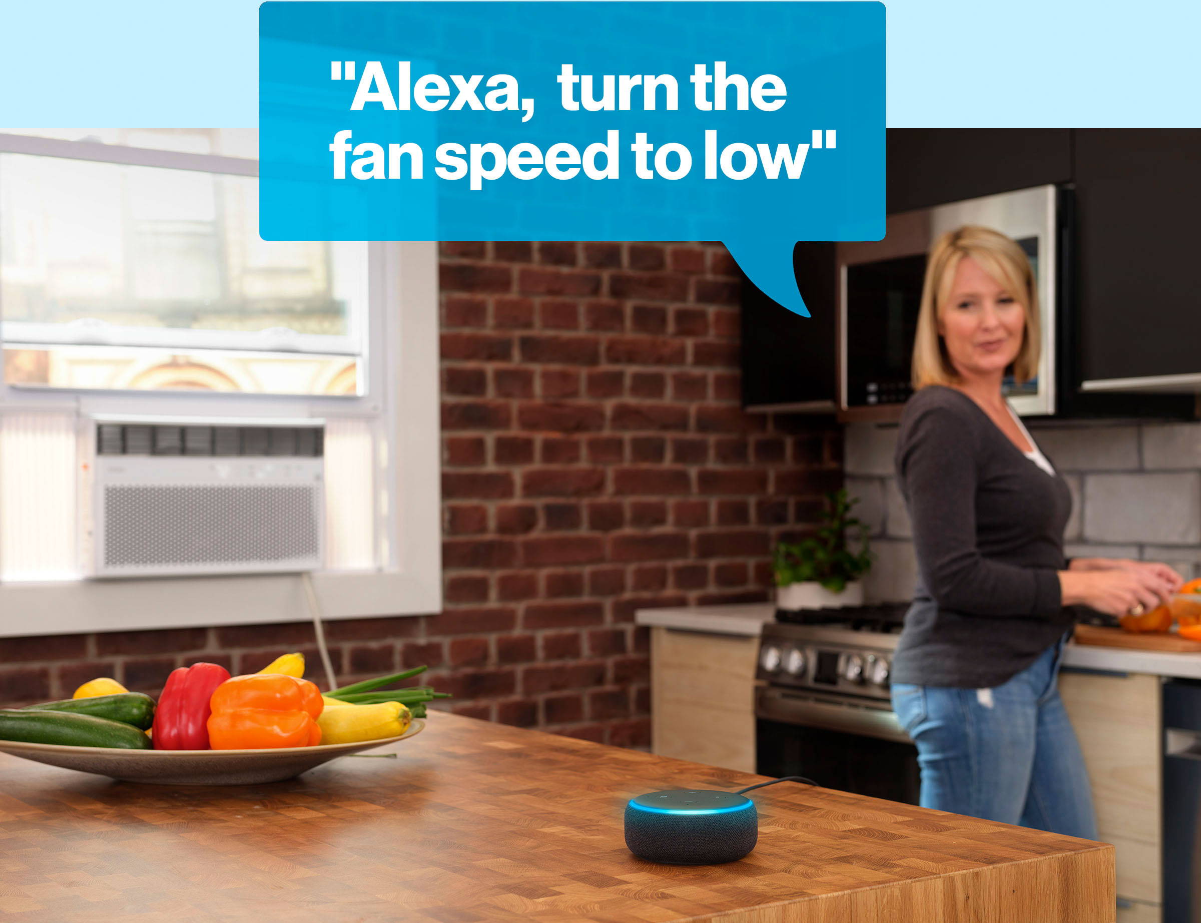 Woman standing at kitchen counter chopping peppers in front of a window air conditioner while asking, Alexa, turn the fan speed to low.