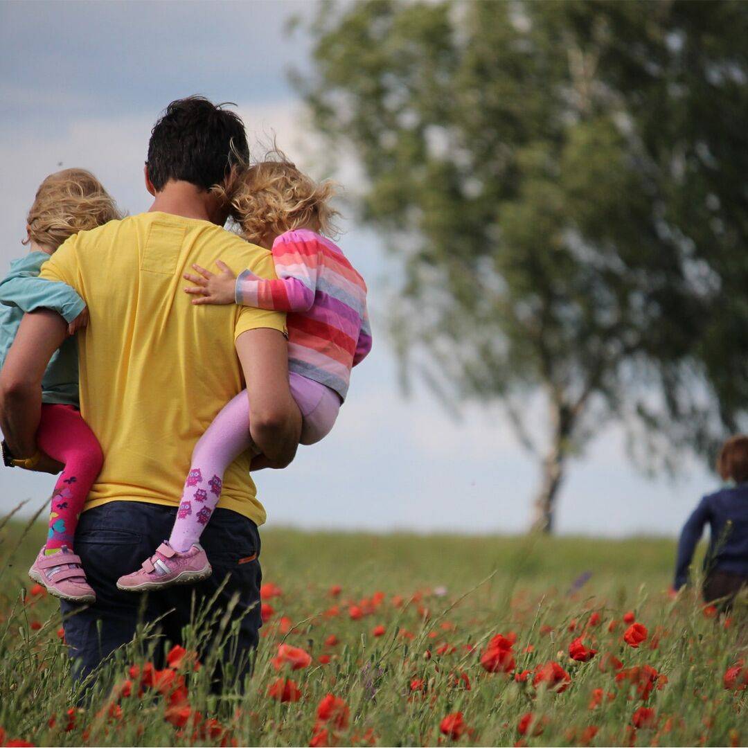 Vater trägt zwei kleine Mädchen durch ein Feld mit langem Gras und roten Mohnblumen, während ihr Bruder vorausläuft