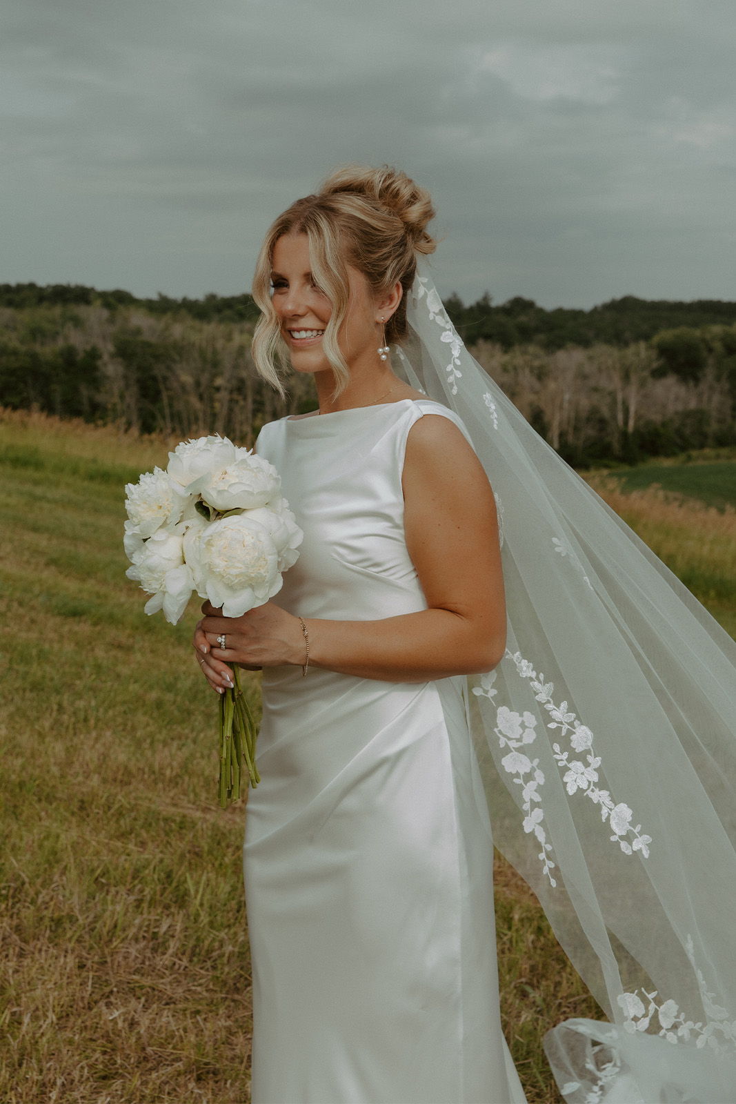 Bride in a wedding dress holding white flowers outdoors, smiling.