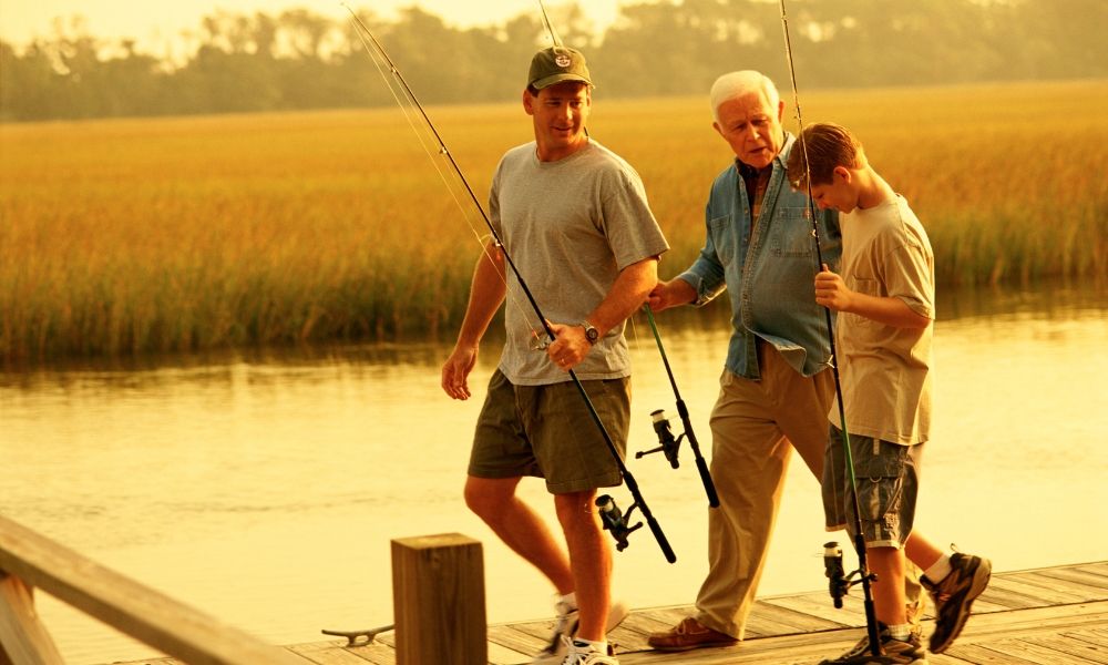 Three men walking by a lake, holding fishing rods at sunset.