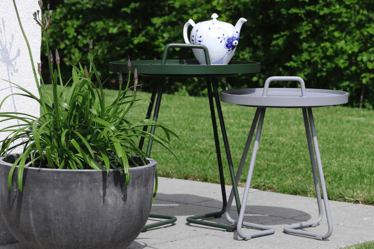 A white teapot on a green side table beside a smaller matching side table in grey with plant in the foreground.