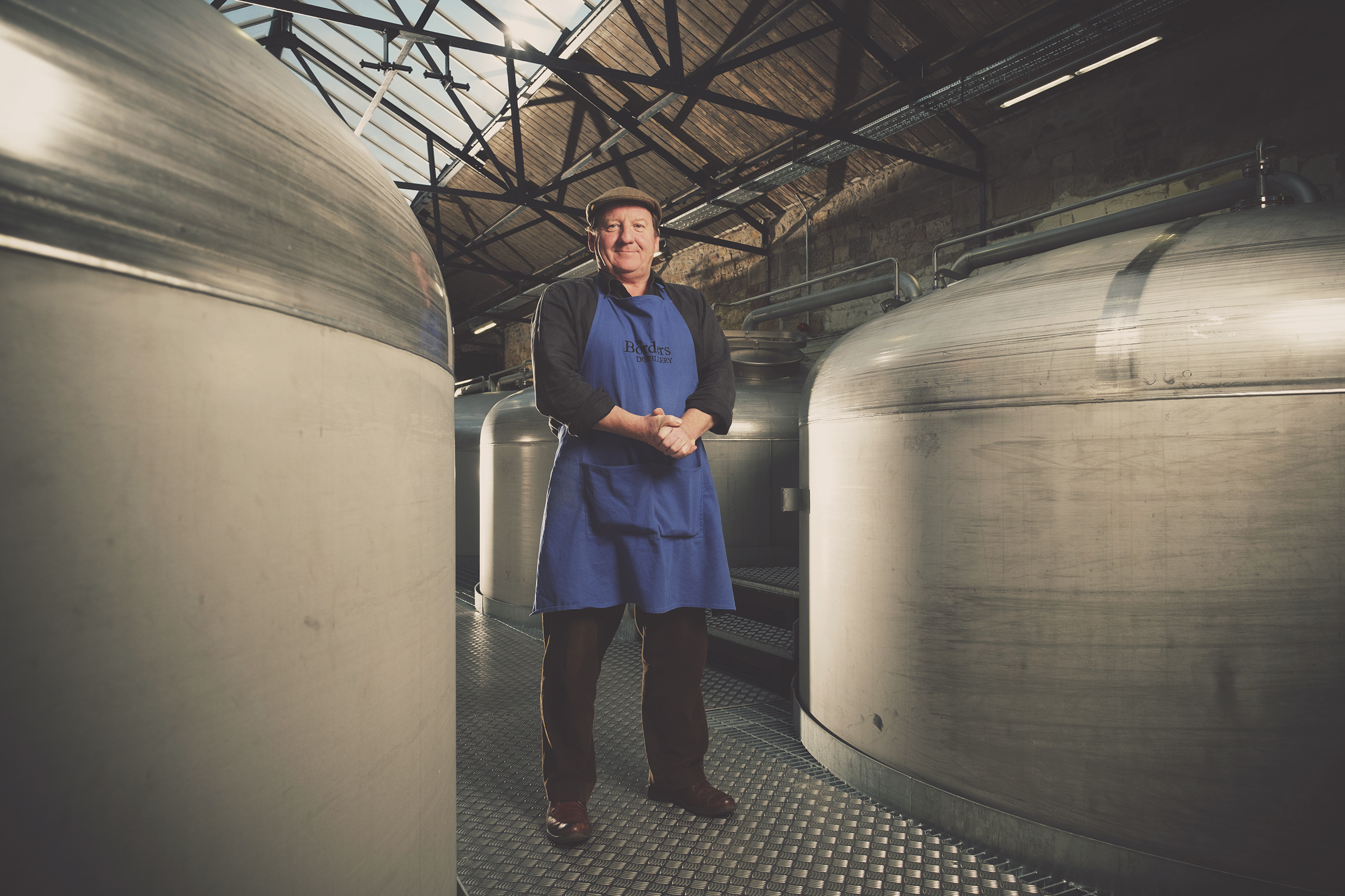 Person in an apron stands between large metallic tanks in an industrial setting.