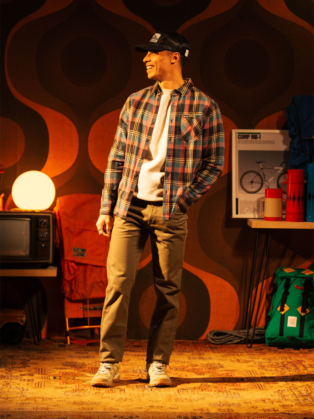 Man wearing a plaid shirt and cap, standing in a retro-themed room.