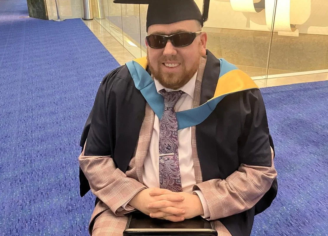 Graduating individual in cap and gown, seated with sunglasses, smiling.