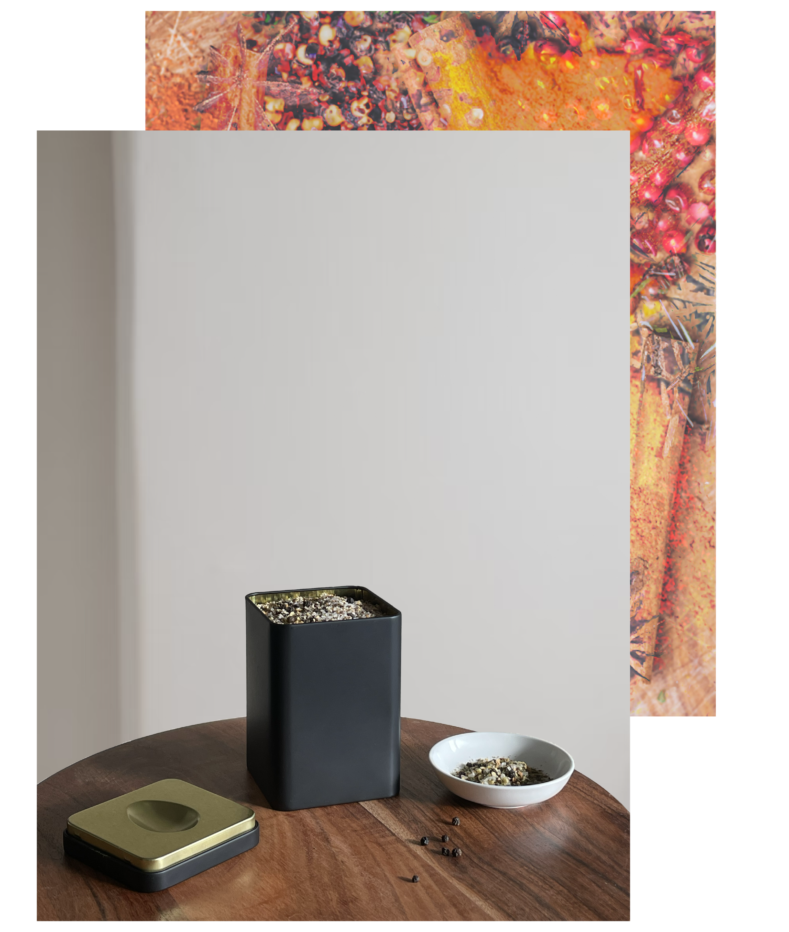 loose leaf tea packaging with holiday spice mix in tin with bowl on the right side on a wooden table the lid of the tin on the left side  loose pepper corn on the table. An image of a stylized mix of holiday spices is in the background.