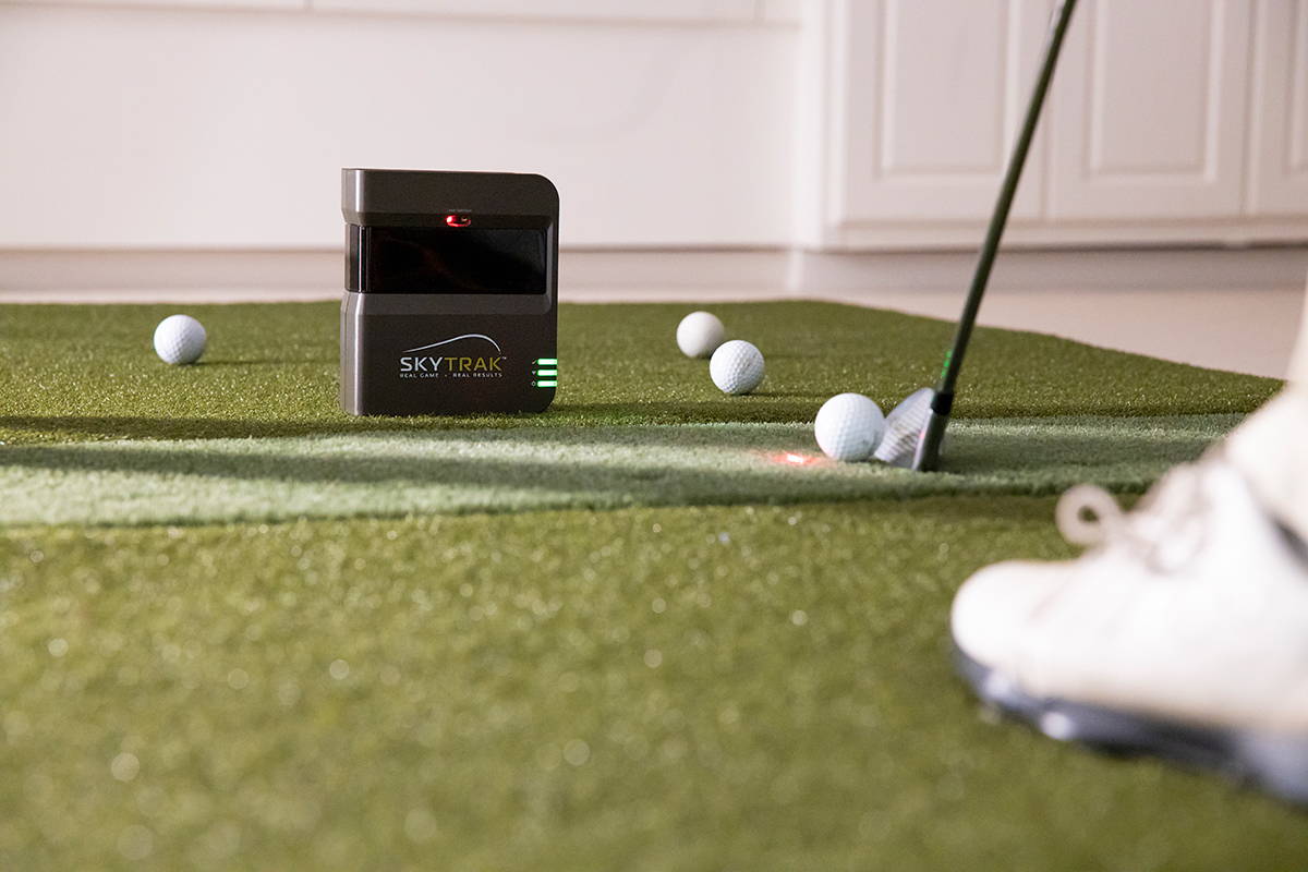 A SkyTrak golf launch monitor unit on a golf mat with several golf balls, a golf club and a golfer's foot in view