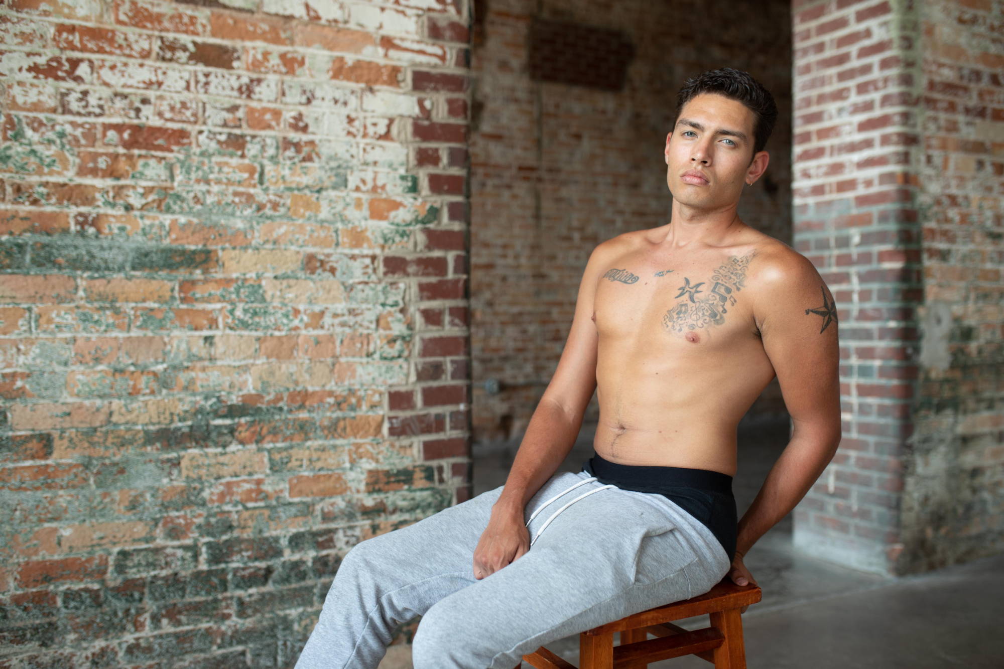 A man in gray sweats and black underwear sits on a wooden stool against a brick wall.