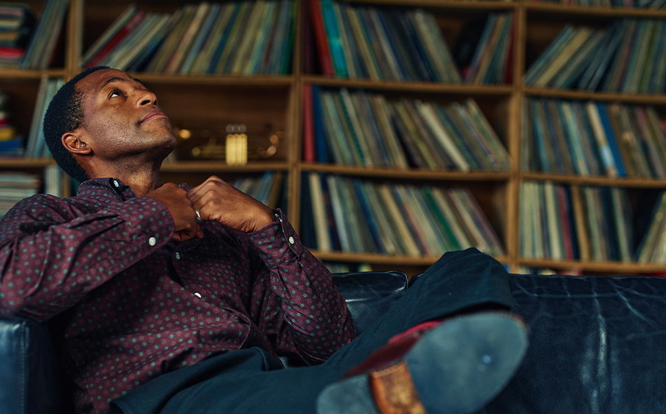 A man sits on a couch, gazing thoughtfully at a record collection.