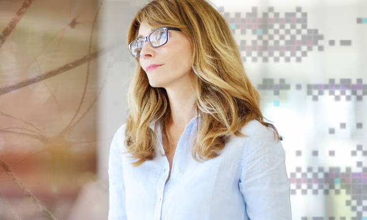 Woman with long hair wearing glasses, standing in a modern environment.