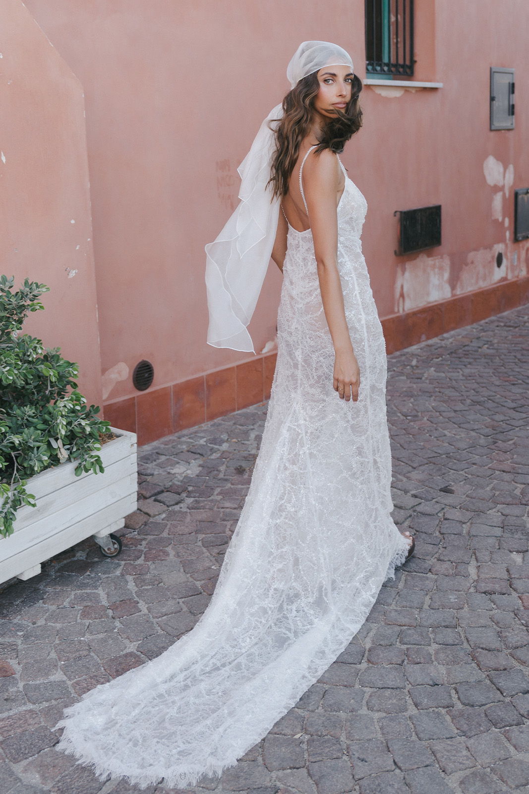 Woman in a lace wedding dress with a veil, standing on cobblestone street.