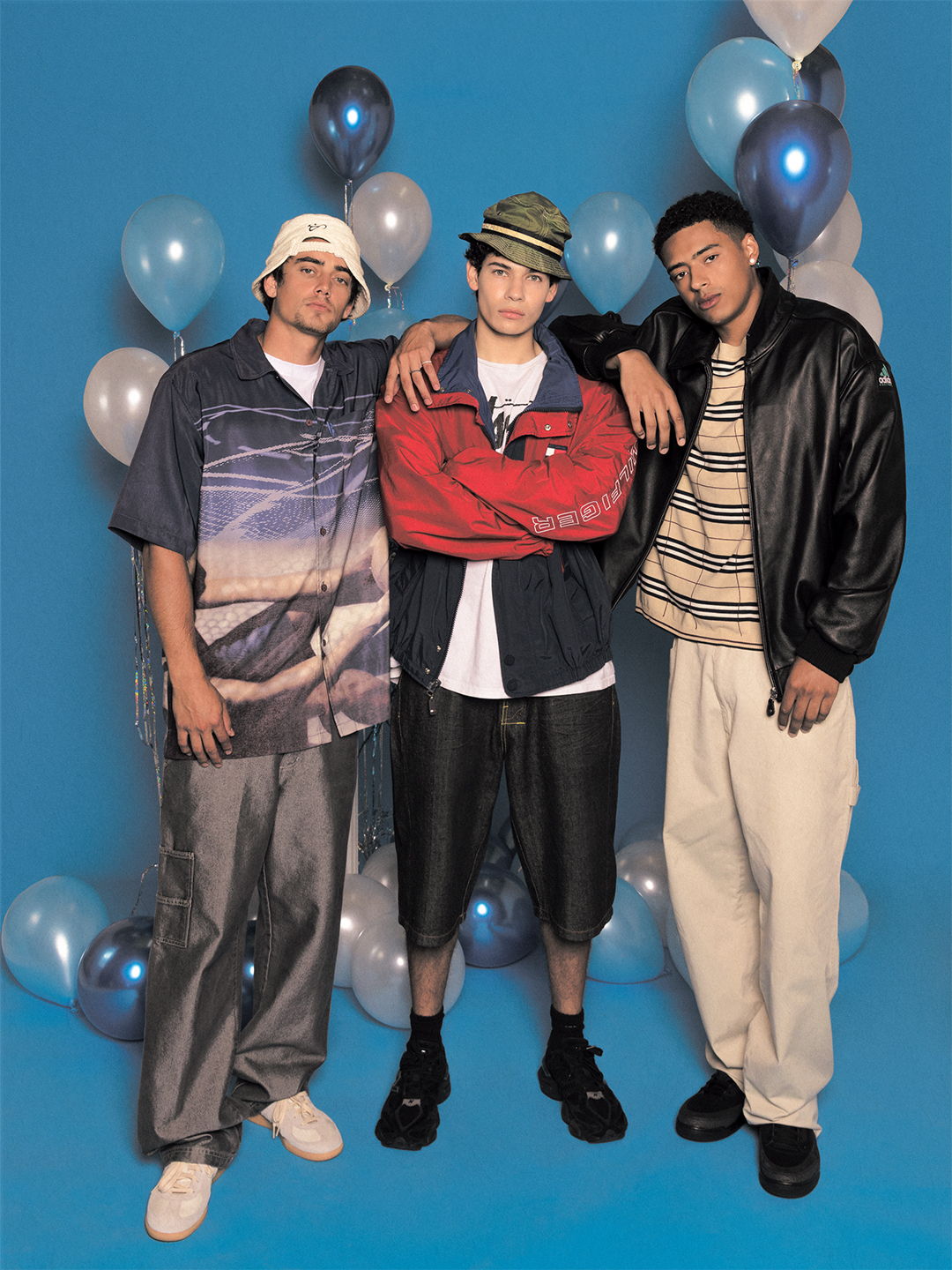 Three young men in casual outfits pose against a blue background with balloons.