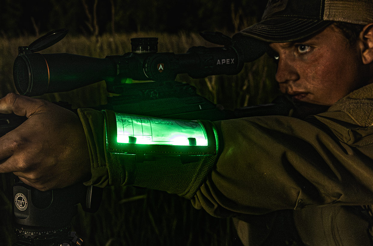 Person aiming a rifle with a green illuminated device in low light.