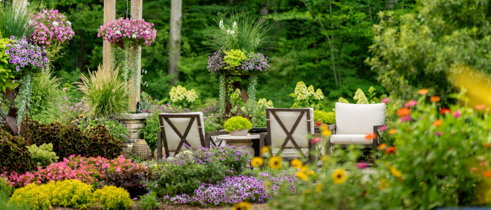 Bright, lush garden patio with a wooden pergola, colorful flowering plants, and empty outdoor seating surrounded by greenery and trees.