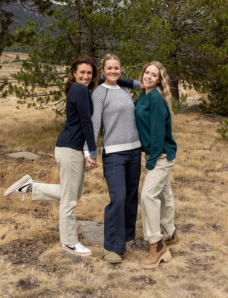 Three women in casual outfits pose outdoors among trees and grass.