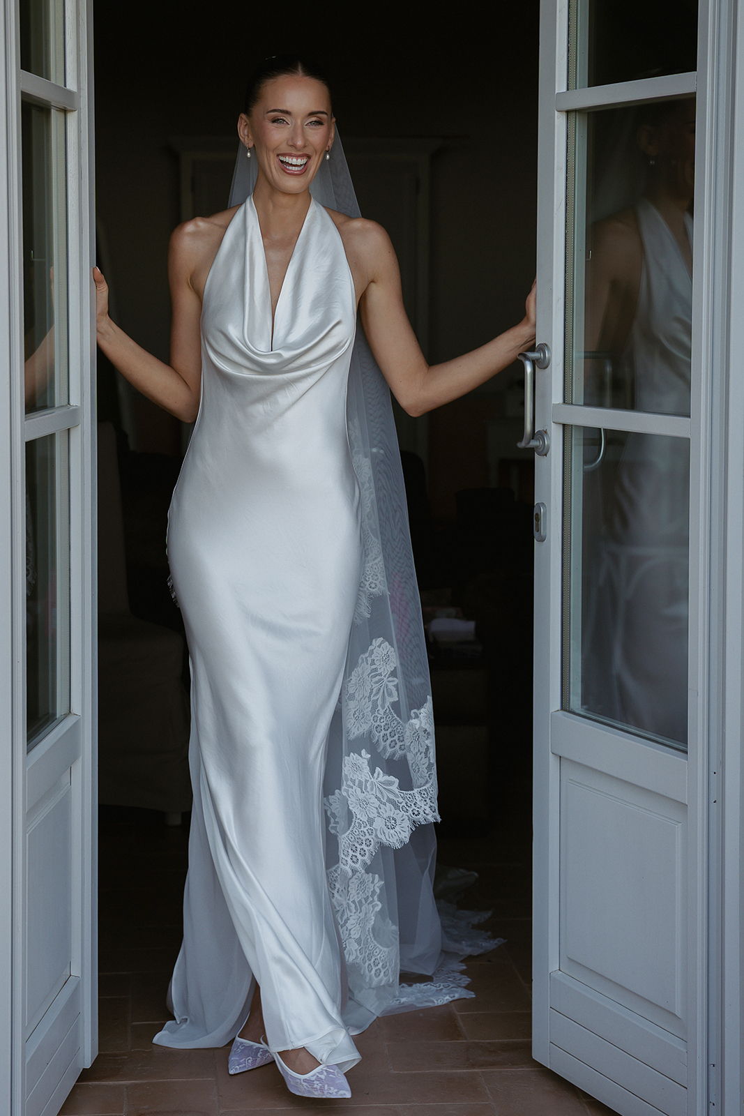 Smiling woman in white wedding dress and veil standing in doorway.