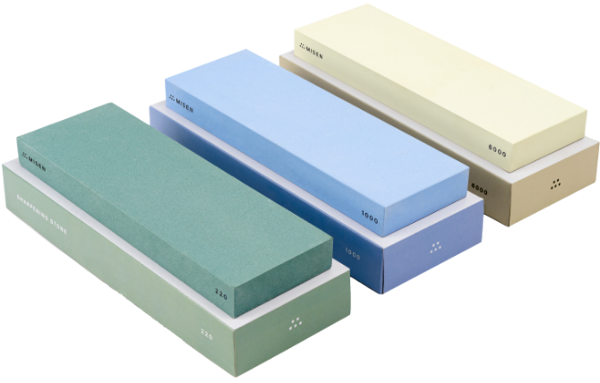 A green Misen 220-grit sharpening stone, a blue Misen 1000-grit sharpening stone, and a cream Misen 6000-grit sharpening stone laid side by side atop their storage boxes.