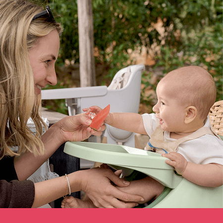 A mother feeding her smiling baby in a high chair outdoors.