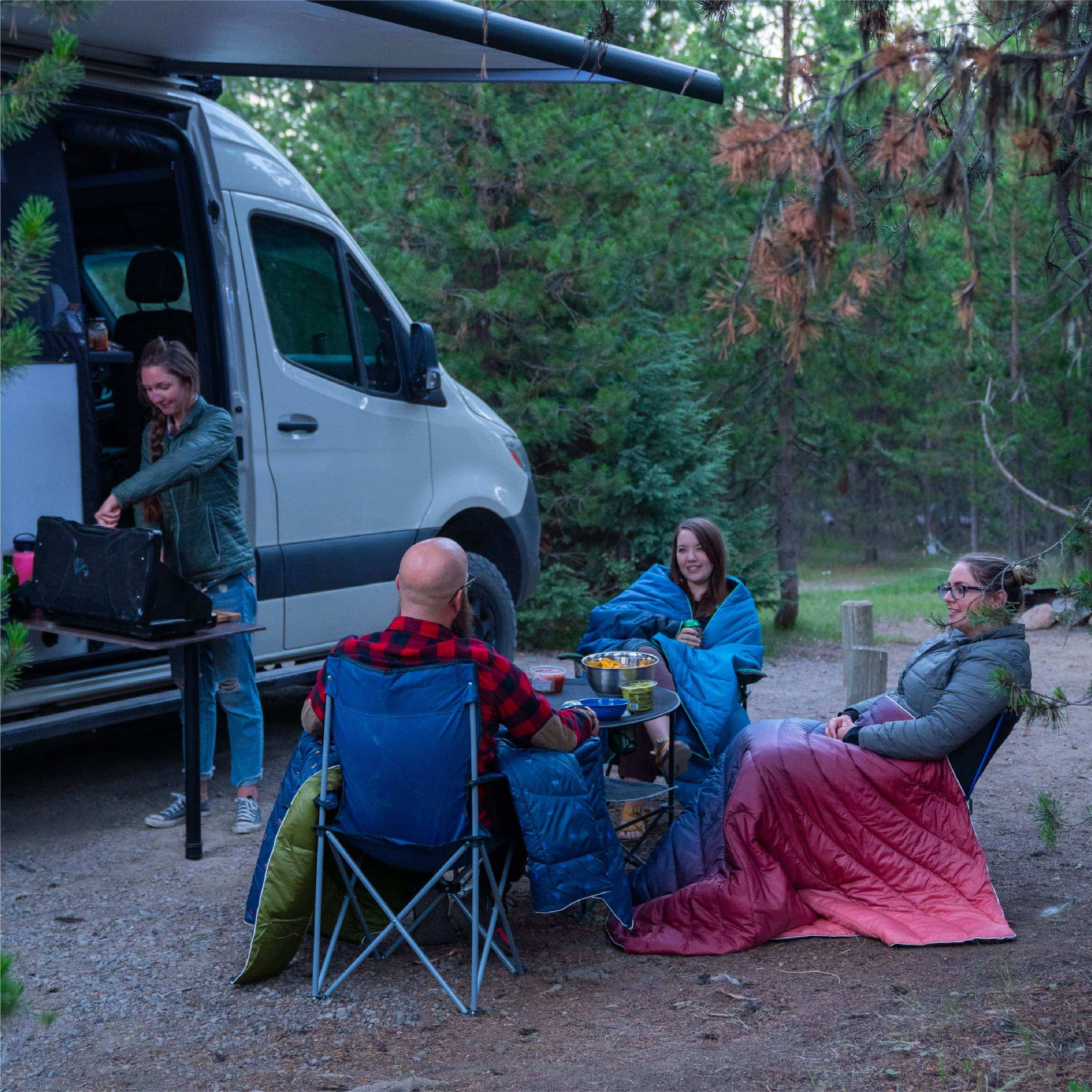 Group of Four Men and Women Having A Picnic Outside Of Camper Van While Wrapped In Original Puffy Blankets