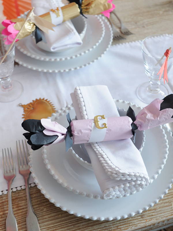 A stack of pearl white china with a homemade pink and black tissue paper cracker on top, finished with a gold initial.