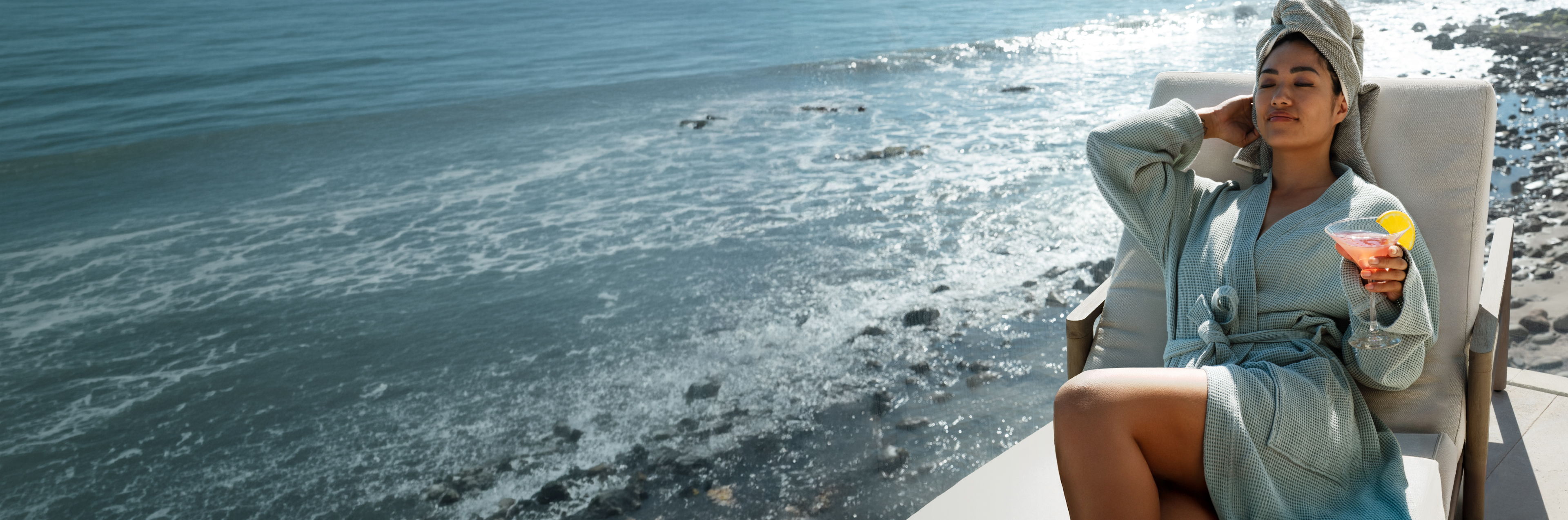 a woman wearing a teal waffle knit robe sitting on a deck at a beachfront house. 