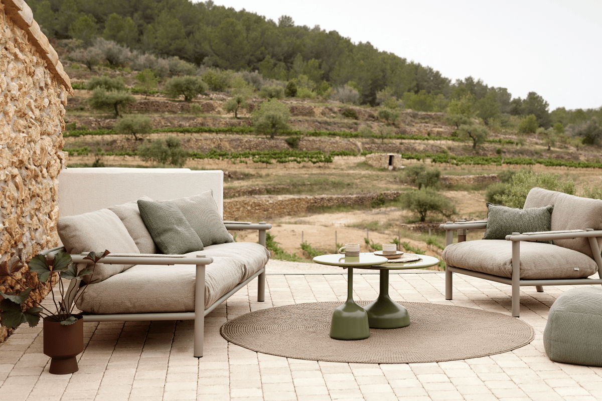 Outdoor lounge arrangement with two low sofas, green pedestal side tables, and a round woven outdoor rug on a stone paver terrace.