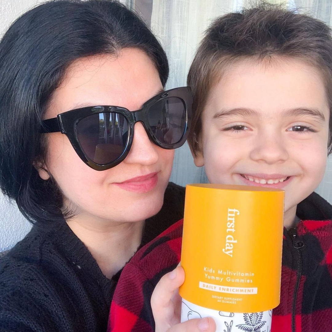 A mom wearing shades smiling with her son who is holding up a First Day vitamins bottle