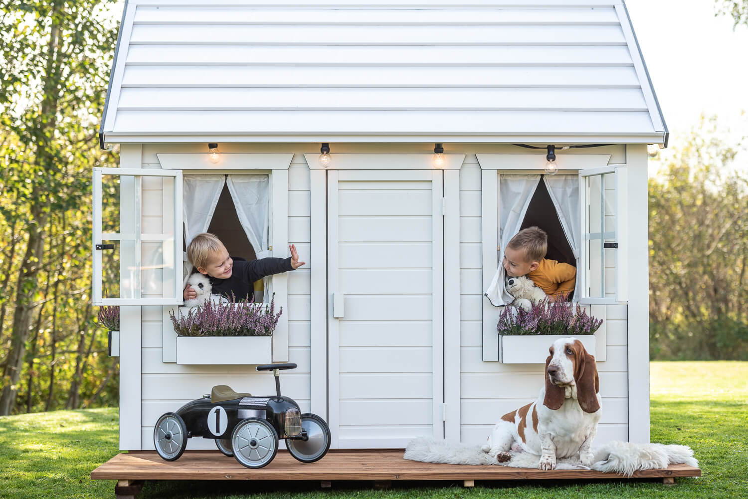 Wooden Playhouse in white color with two boys playing and a car and a dog in front by WholeWoodPlayhouses