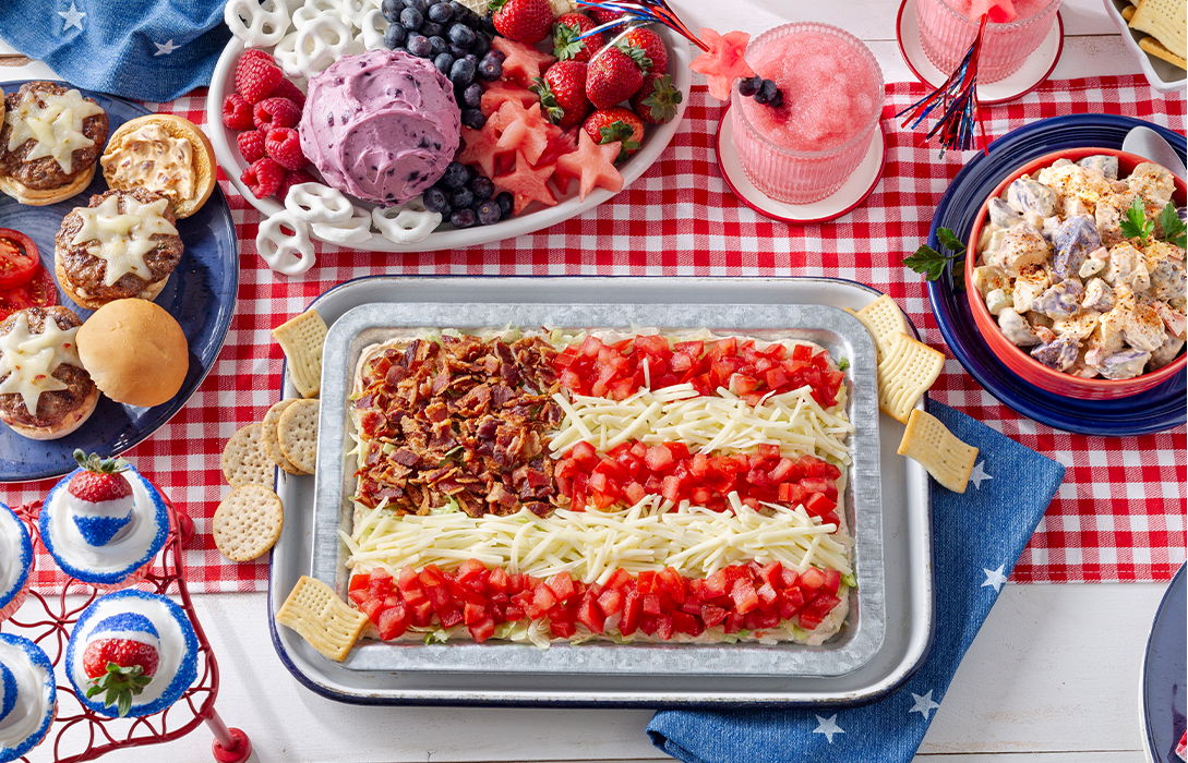 Red, white & blue food spread featuring bacon, cheese and tomato pizza along with cheeseburger sliders, pasta salad and cheese ball dessert with fruit