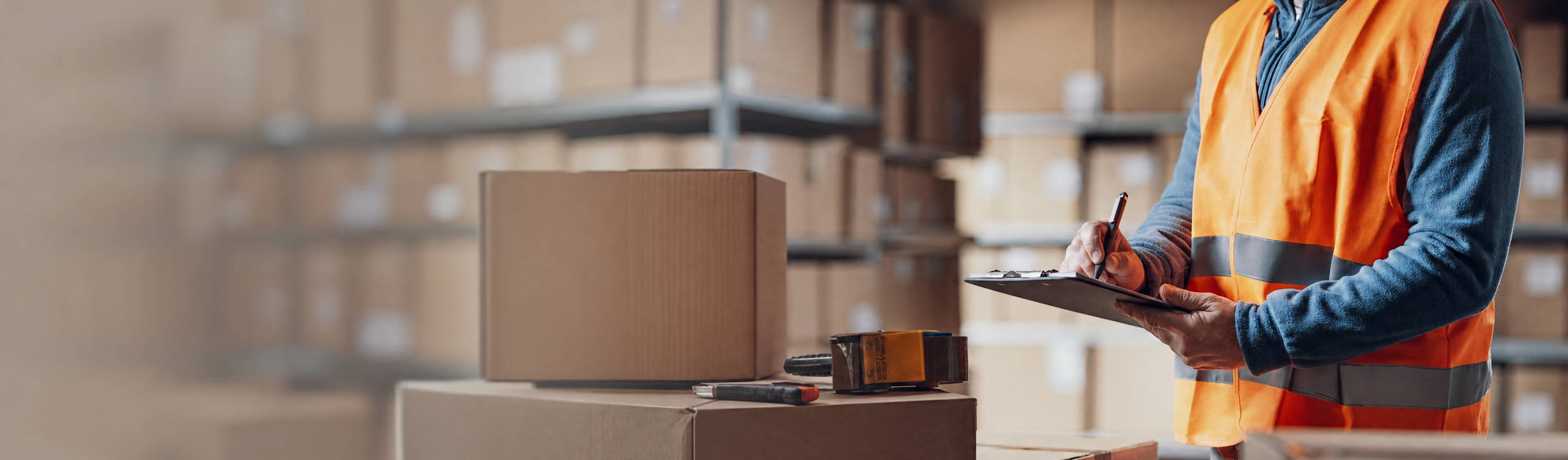 Warehouse worker in safety vest checking boxes with clipboard.