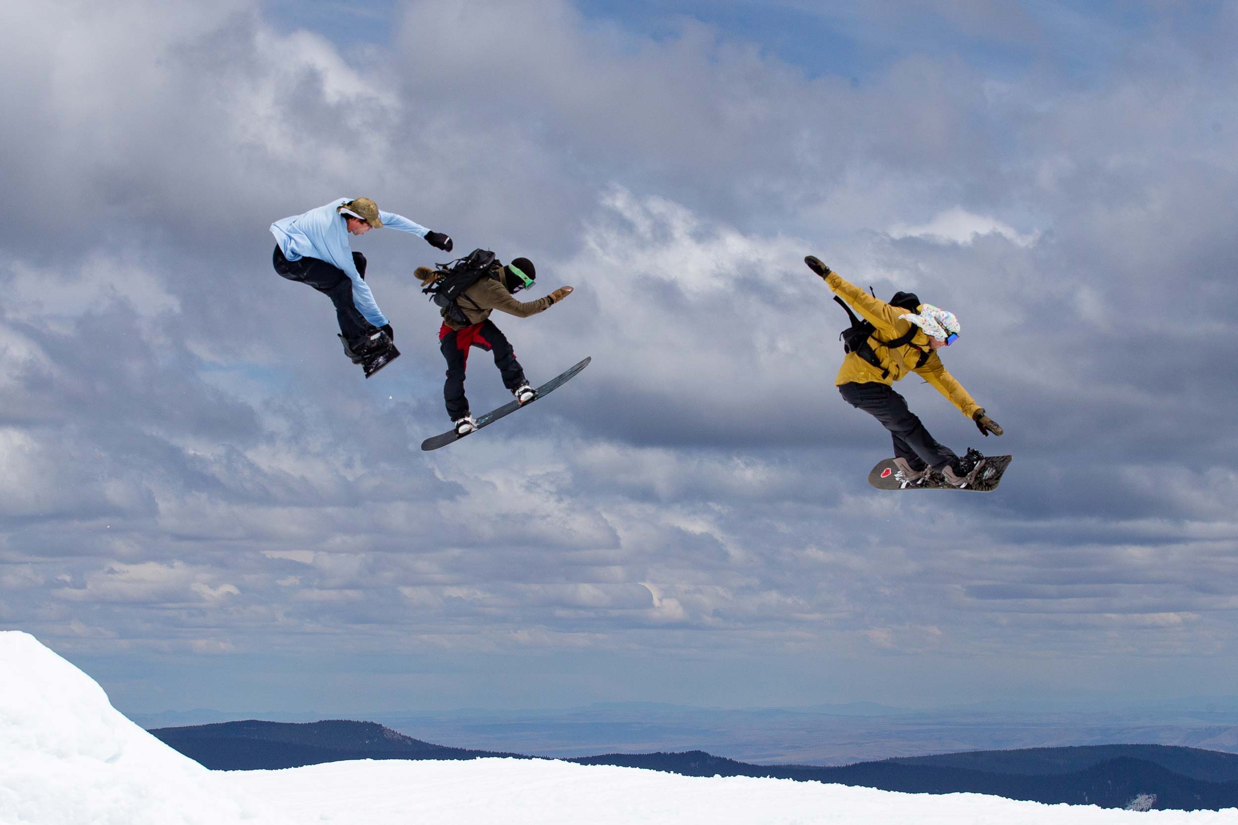 Three snowboarders performing jumps against a cloudy sky.