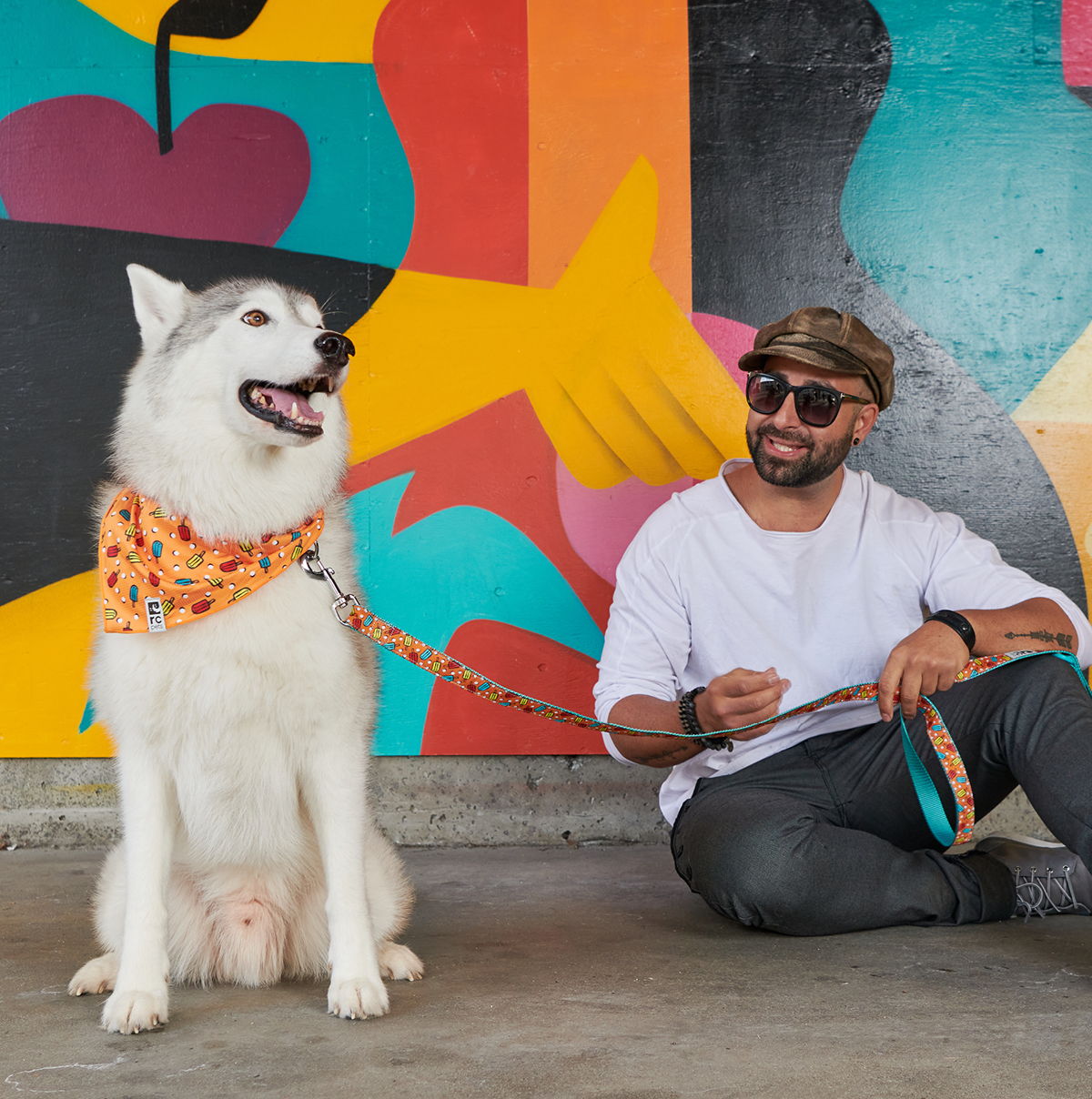 Person sitting next to a dog in front of a colorful mural.