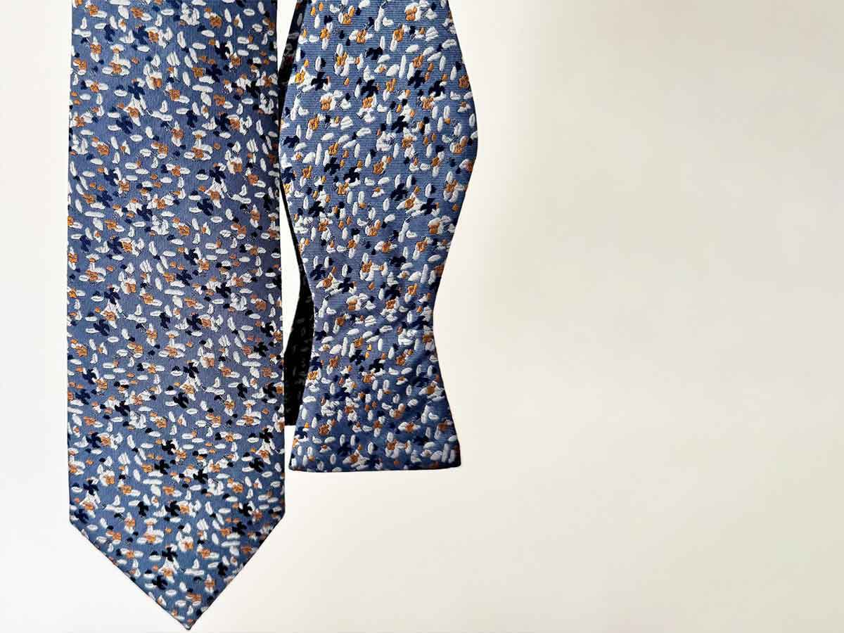 A dusty blue pattern necktie and bow tie, hanging in front of a white background