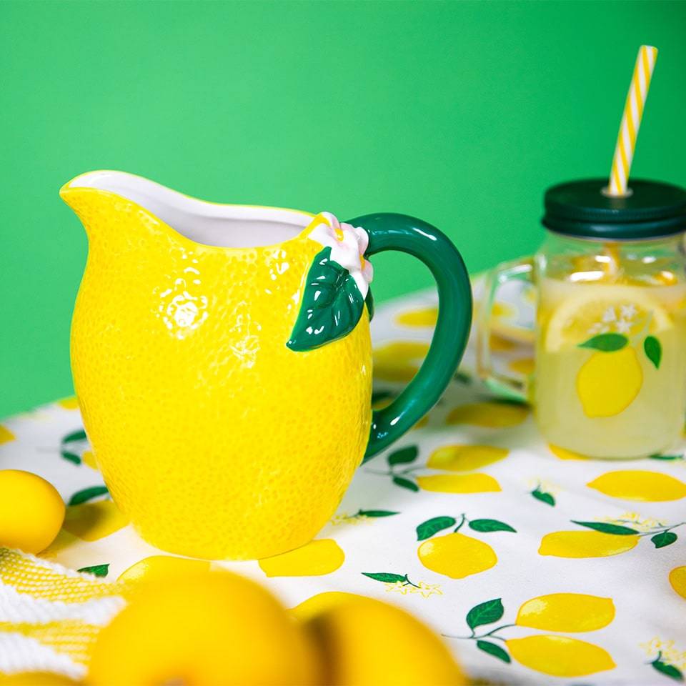 Close-up of a lemon-themed pitcher with a detailed lemon texture and leaf-shaped embellishments, next to a glass with a lemon slice and a striped straw, set on a lemon-print tablecloth.