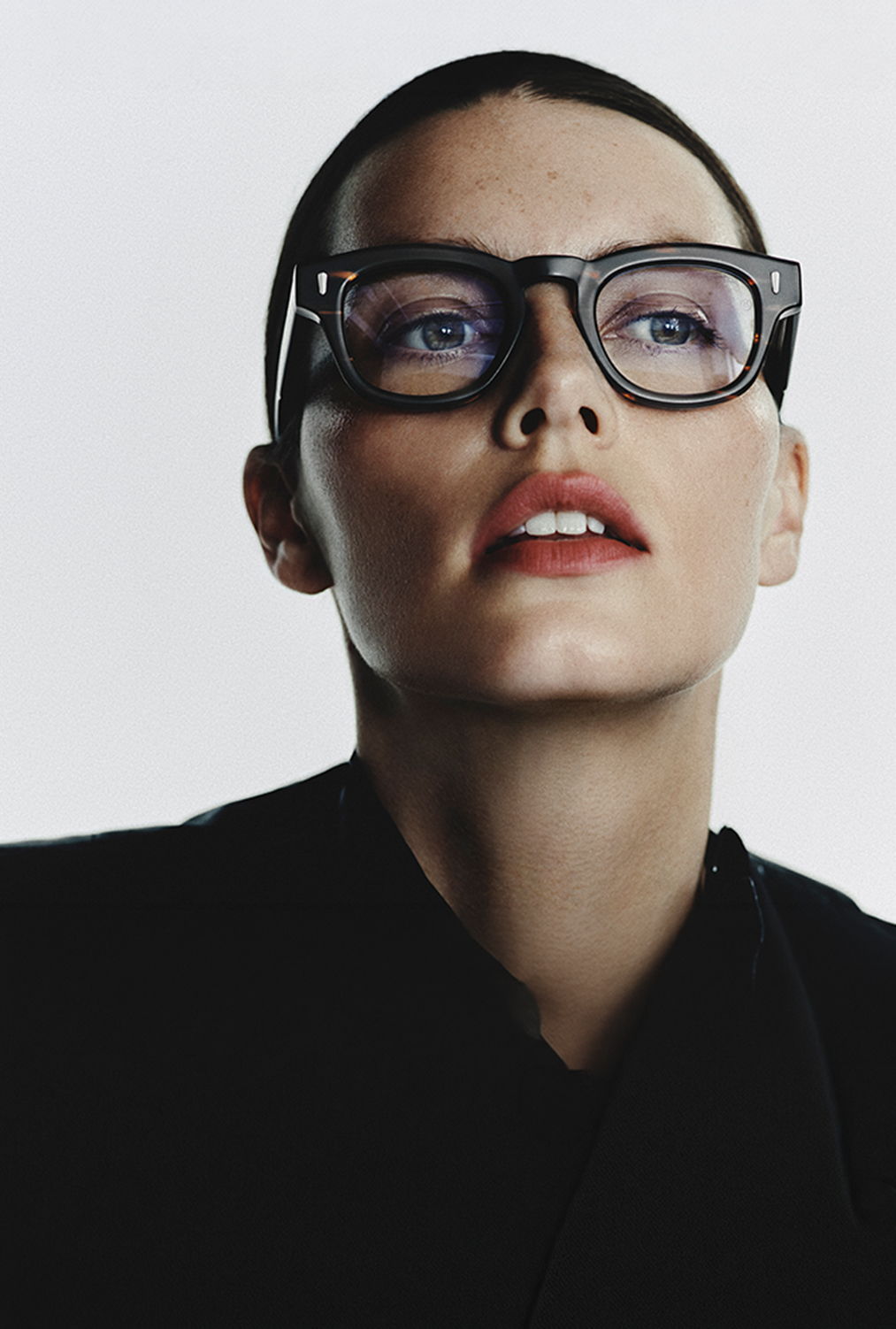 Person with short hair and glasses, wearing a black outfit, looking upward.